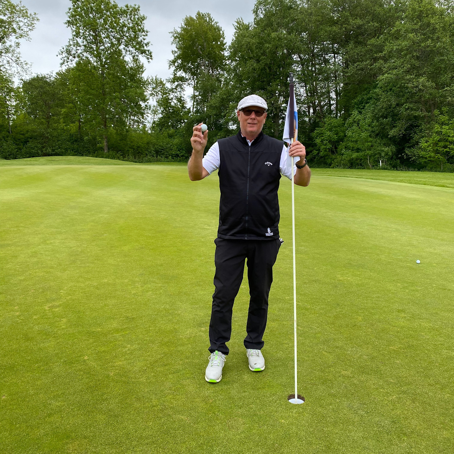 Man holder en golfbold og står ved siden af flagstangen på en golfgreen, omgivet af grønne træer.