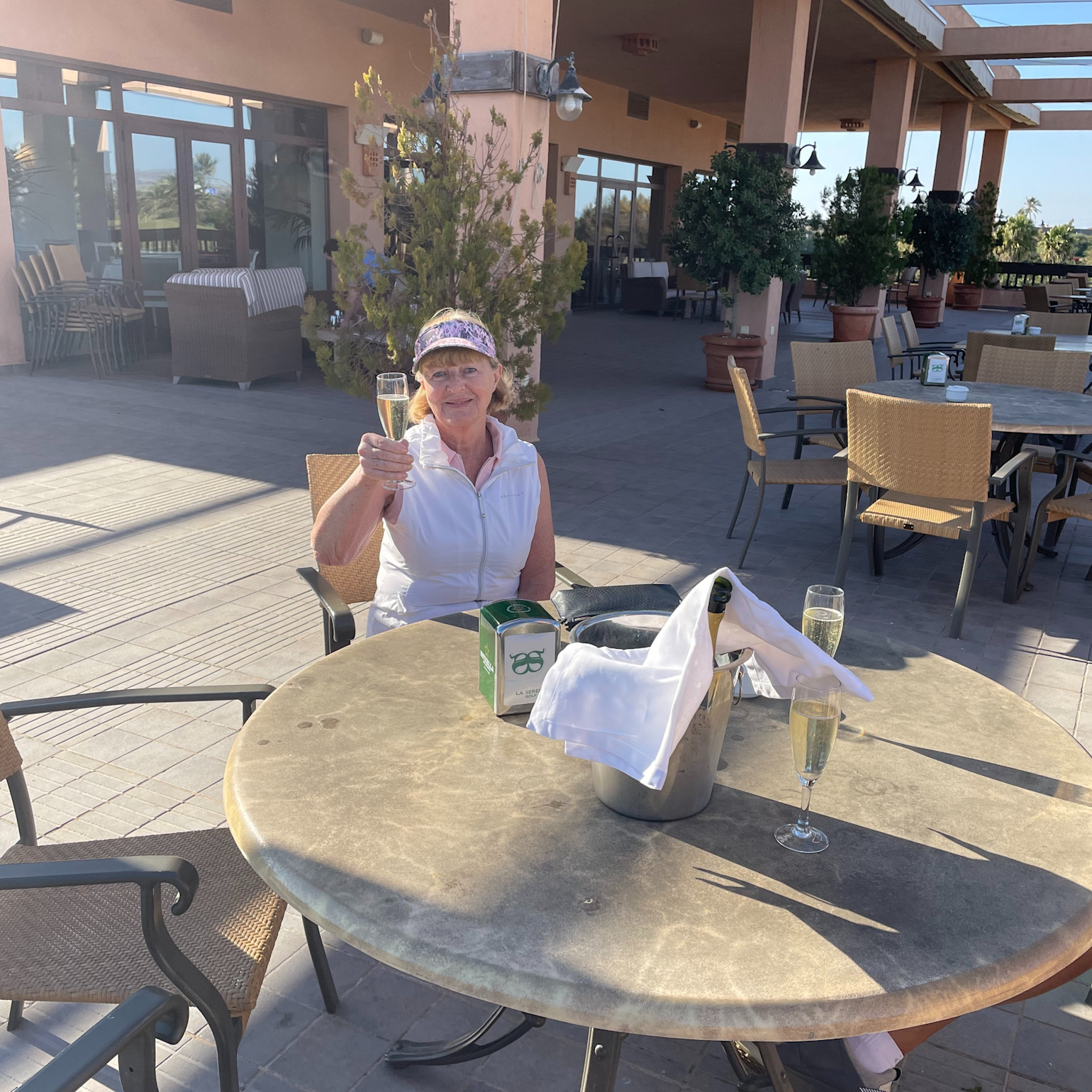 Kvinde i hvid vest sidder ved et rundt bord udendørs, holder et glas bobler op, med champagneflaske og glas på bordet.