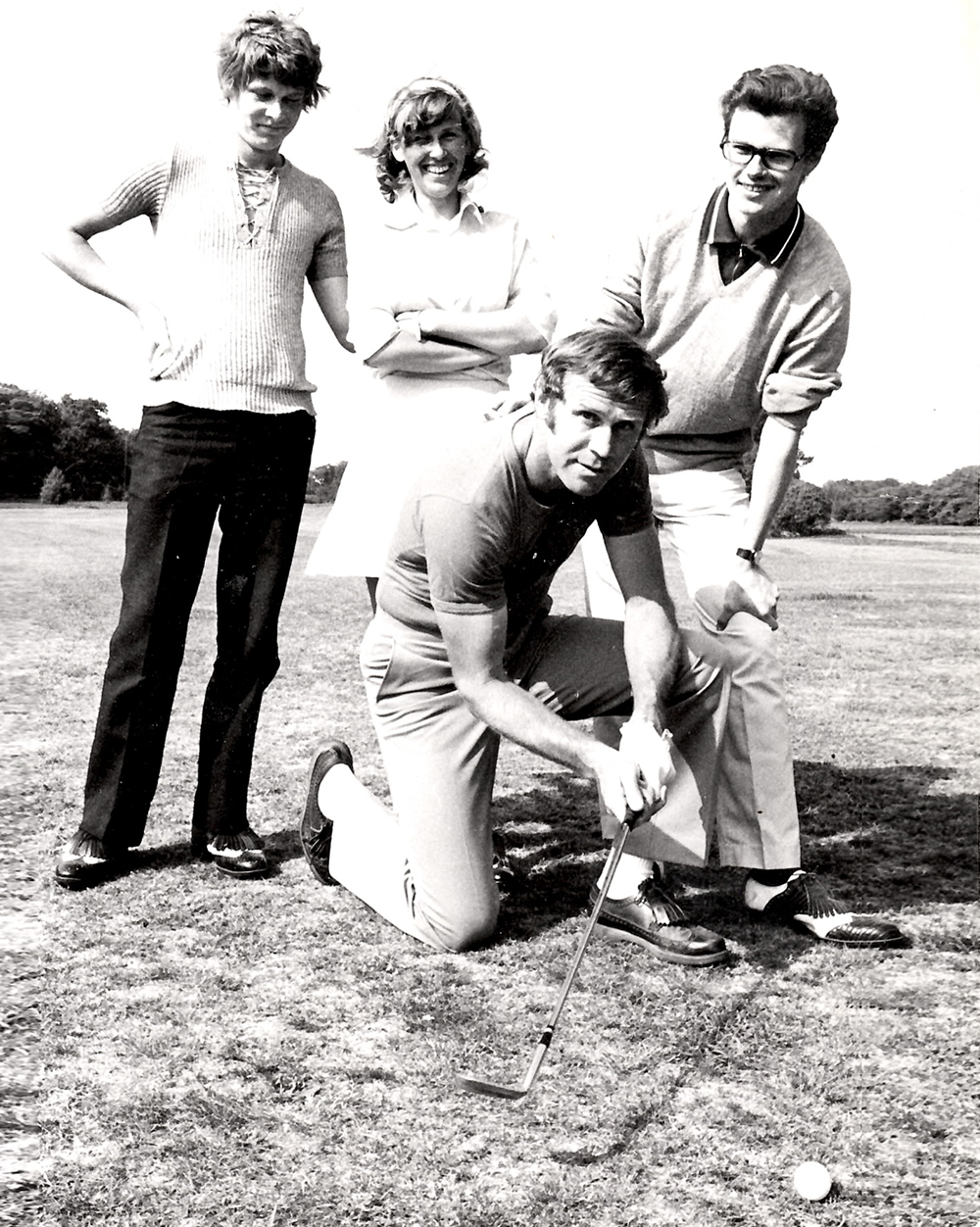 Fire personer poserer på en golfbane; en mand knæler og sigter med en golfkølle mod en golfbold, mens de andre står bagved og smiler.