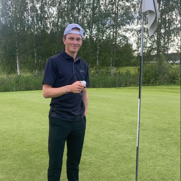 A young man in golf attire is holding a golf ball and standing next to a flagstick on a golf green, with a forest in the background.