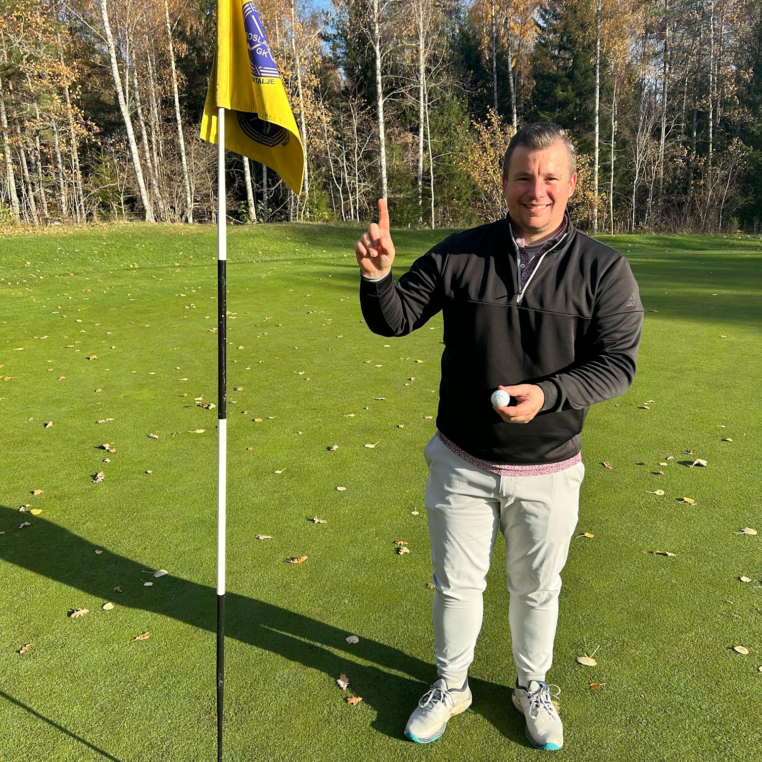 Man håller en golfboll och pekar uppåt med ett finger bredvid en flaggstång på en golfbana omgiven av träd i höstmiljö.