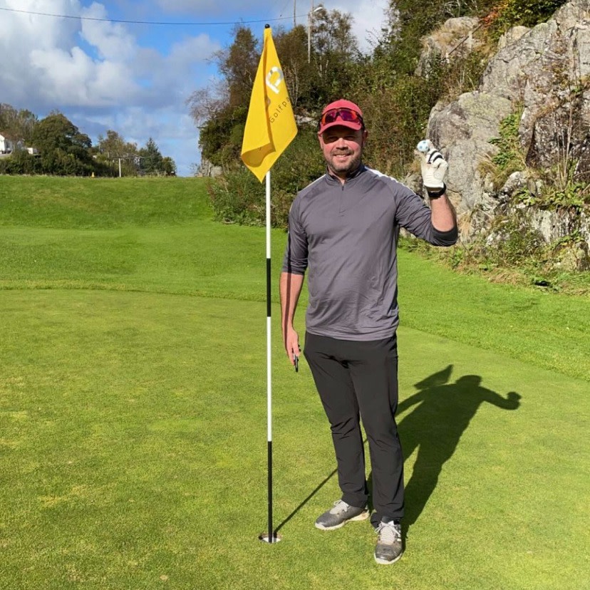 En mand i golfbeklædning står ved siden af en flagstang på en golfgreen og holder en golfbold op, med solskinsvejr og grøn natur i baggrunden.