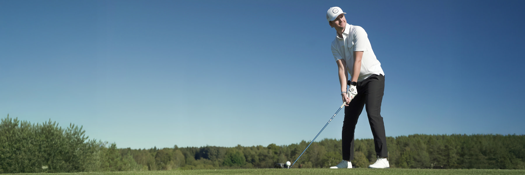 Golfspelare i vit tröja och keps förbereder sig för att slå en boll på en golfbana med skog i bakgrunden.