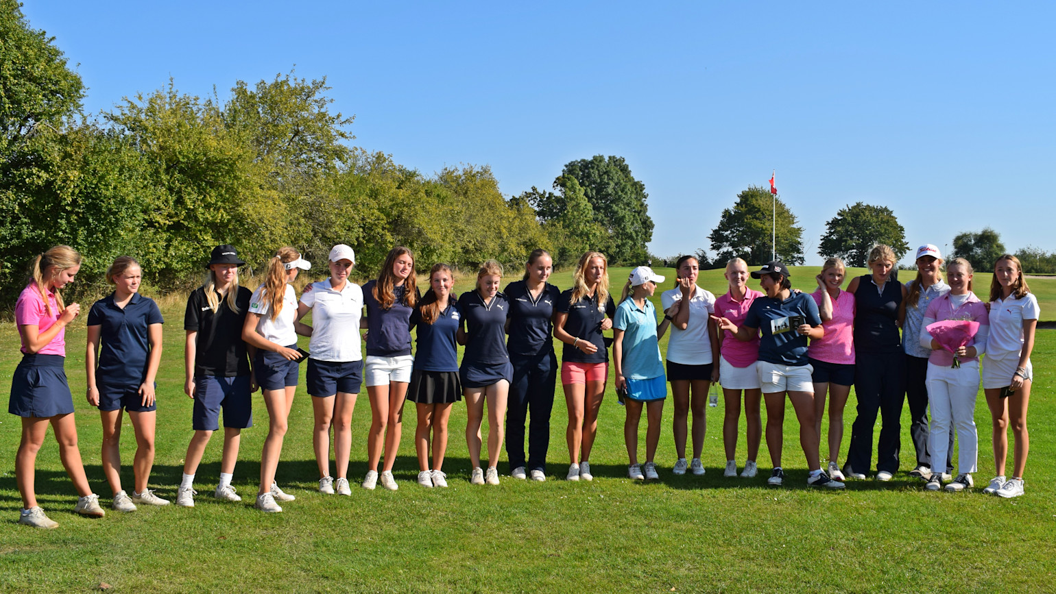 En grupp unga kvinnor står på en golfbana i rad, flera i golfkläder, några håller blommor, soligt väder och grön omgivning.