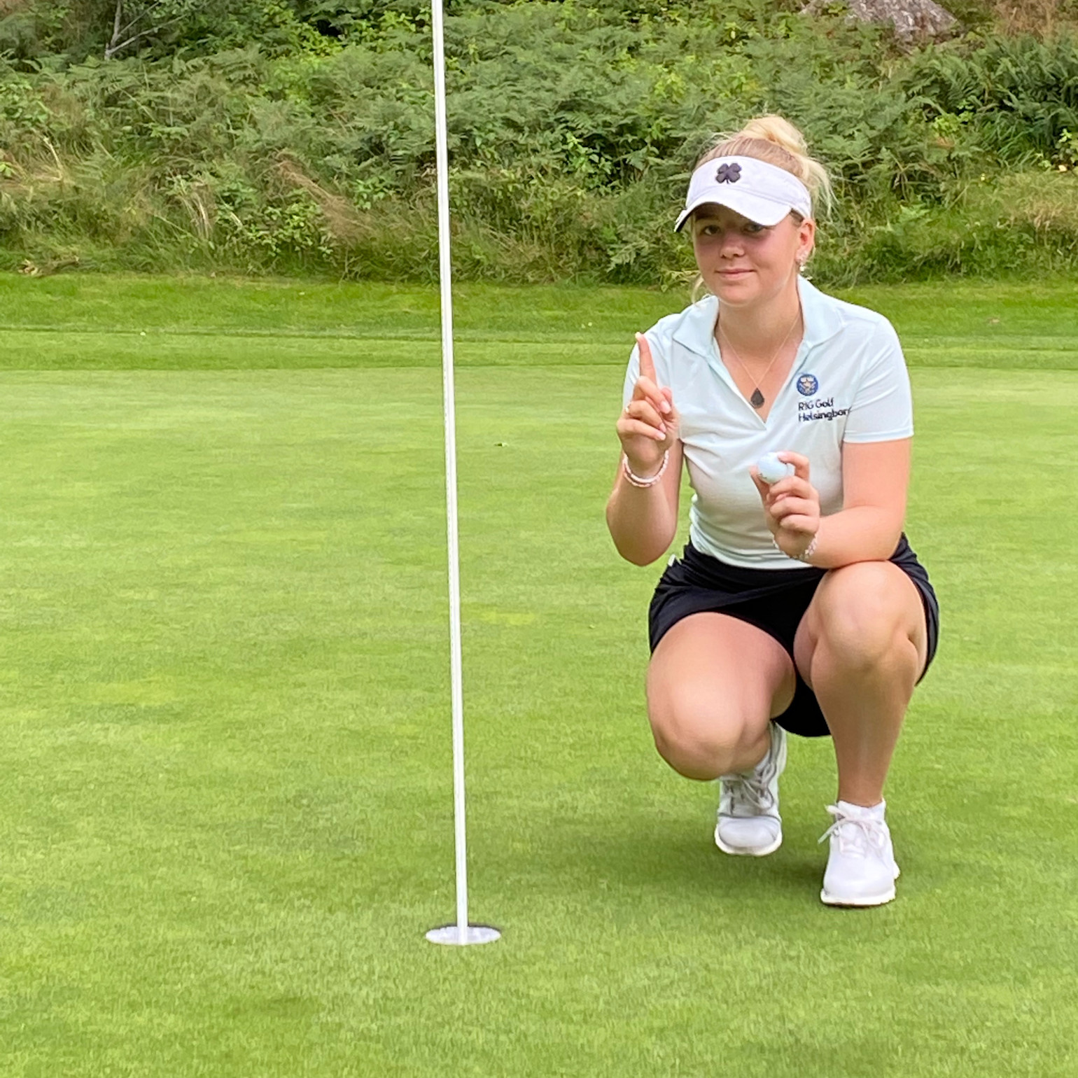 Kvinde i golfbeklædning huker sig ved siden af et golfhul på greenen, holder en finger og en golfbold op, omgivet af græs og træer.