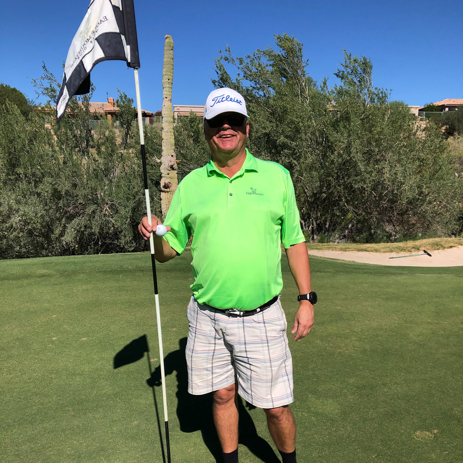 Mand i grøn poloshirt og ternede shorts holder en golfklubsflag på en golfbane under en klar blå himmel.