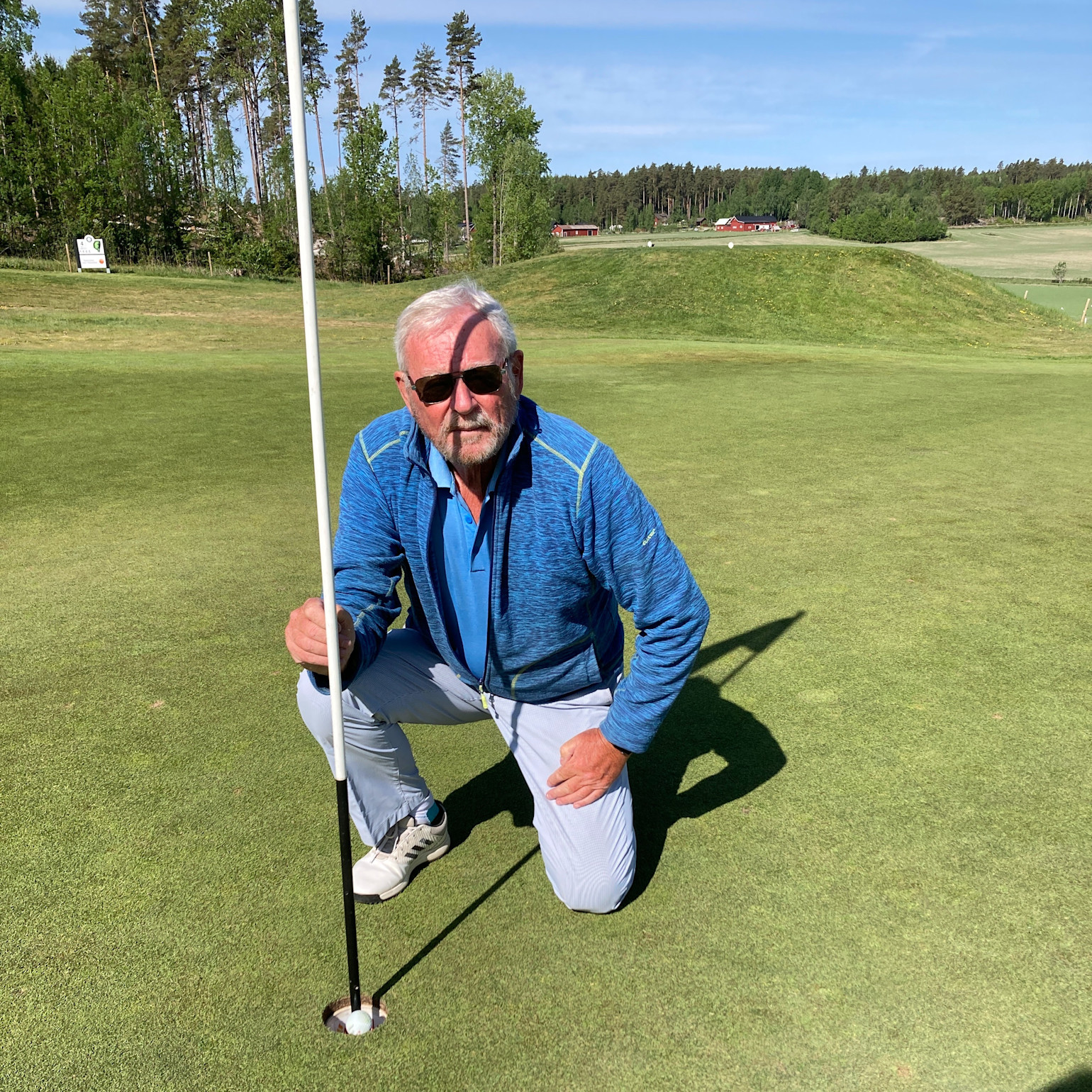 Ældre mand med solbriller og blå jakke huker sig ved et golfhul og holder i flagstangen på en grøn golfbane.