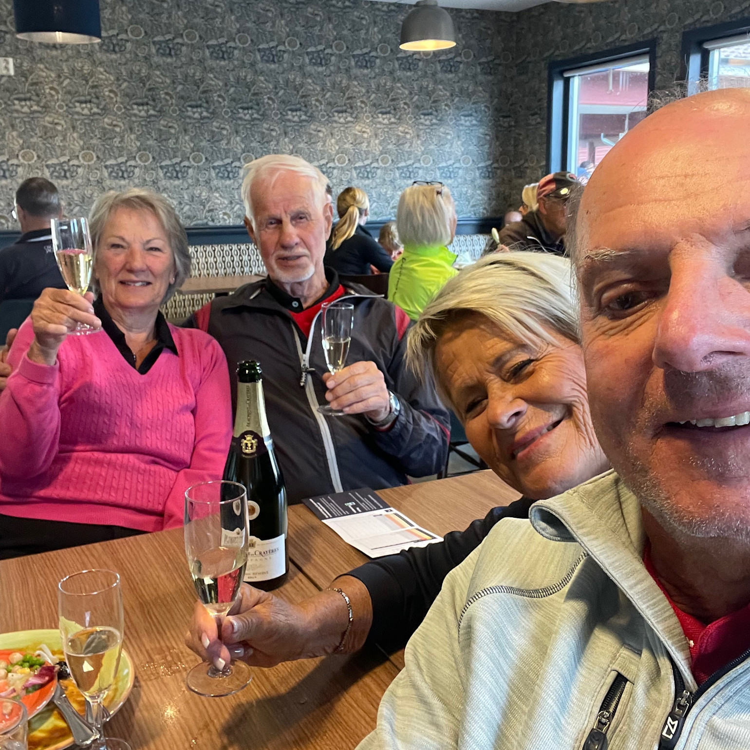 Fire glade personer skåler med champagneglas rundt om et bord på en restaurant, med en flaske bobler og mad foran sig.