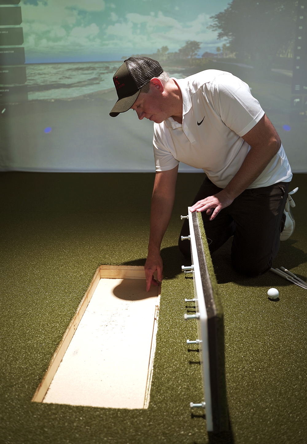 Ein Mann im weißen Poloshirt und mit Mütze untersucht einen geöffneten Abschnitt eines Kunstrasenplatzes in einem Innenraum, neben ihm liegen ein Golfball und Golfschläger.