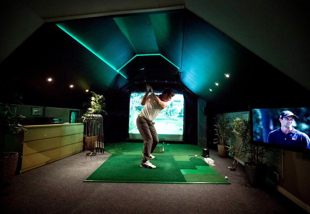 En person svingar en golfklubba i en inomhusgolfstudio med konstgräs, växter och en TV-skärm som visar en golfspelare.