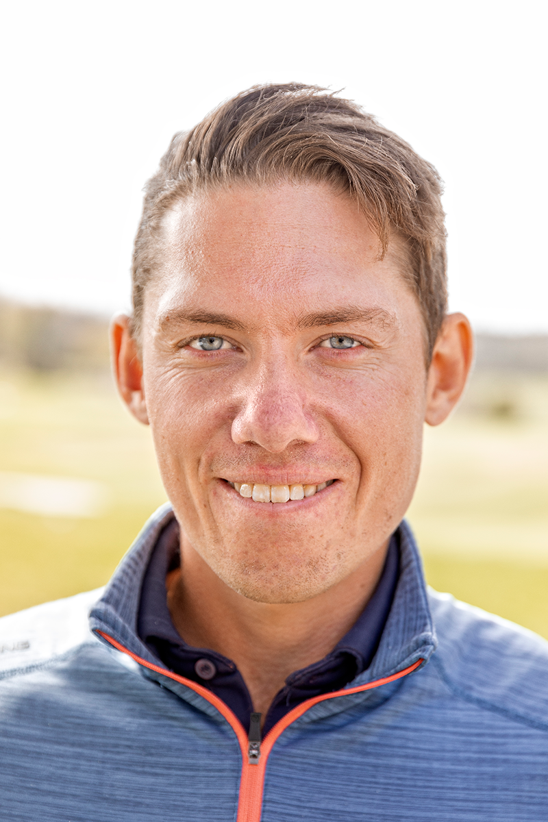 Man ler mot kameran utomhus, klädd i blå jacka med dragkedja, suddig bakgrund med gräs och ljus himmel.