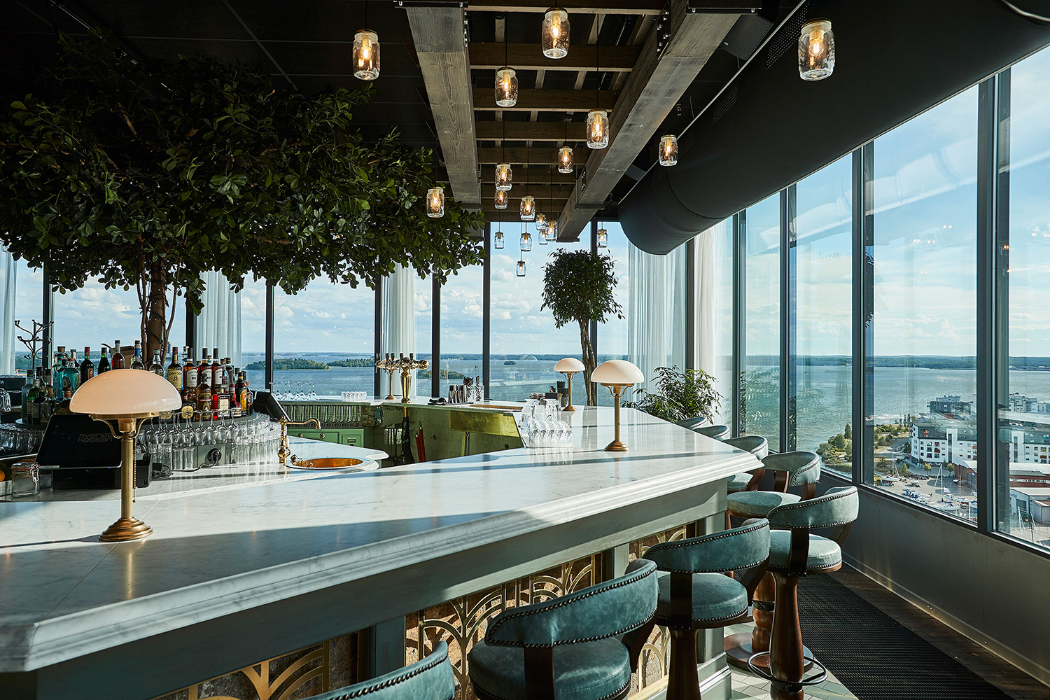Marmorbardisk med gröna barstolar, flaskor och lampor, stora fönster med utsikt över stad och vatten under dagsljus.