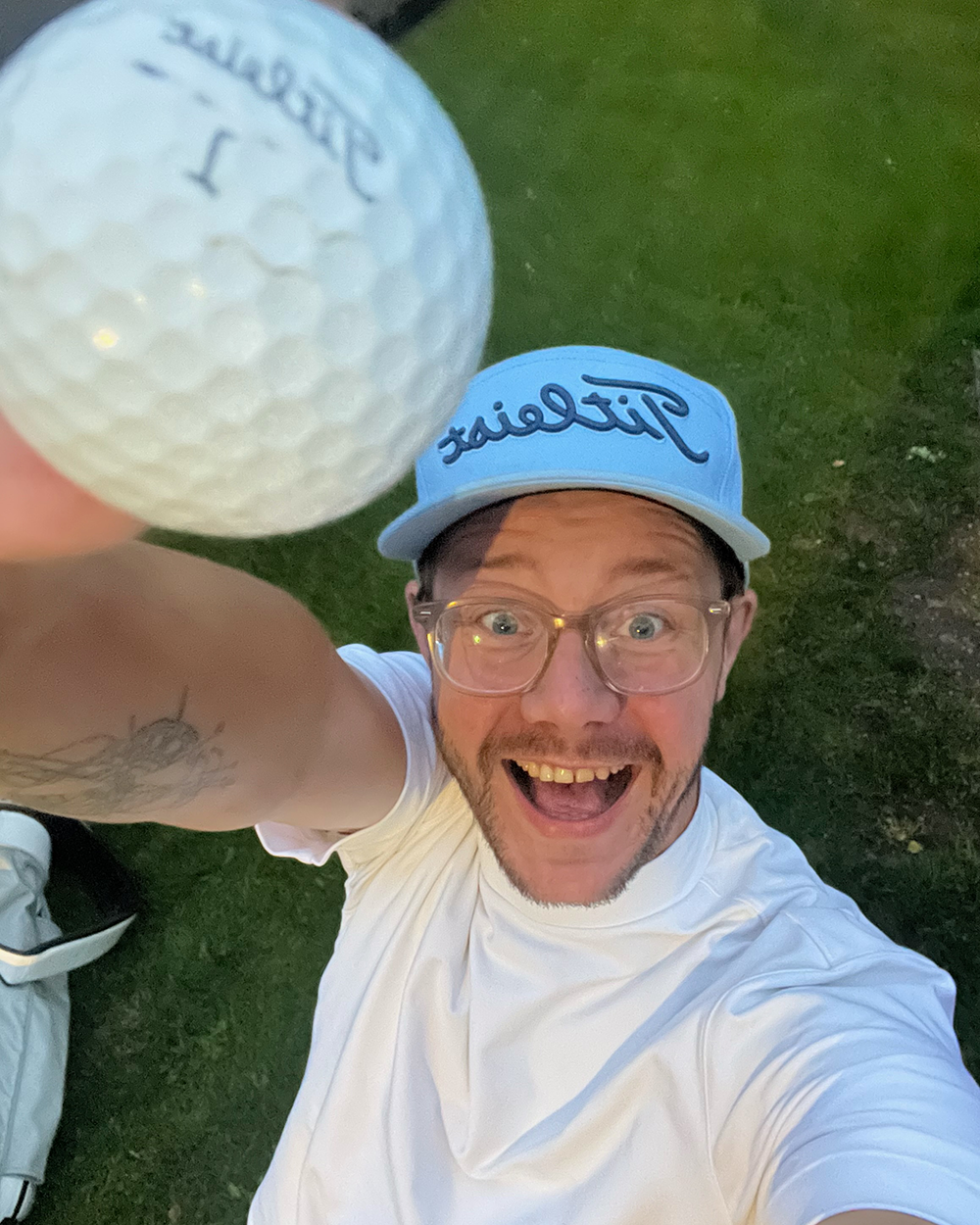 Leende man i glasögon och keps håller upp en golfboll med texten "Titleist" utomhus på en gräsmatta.