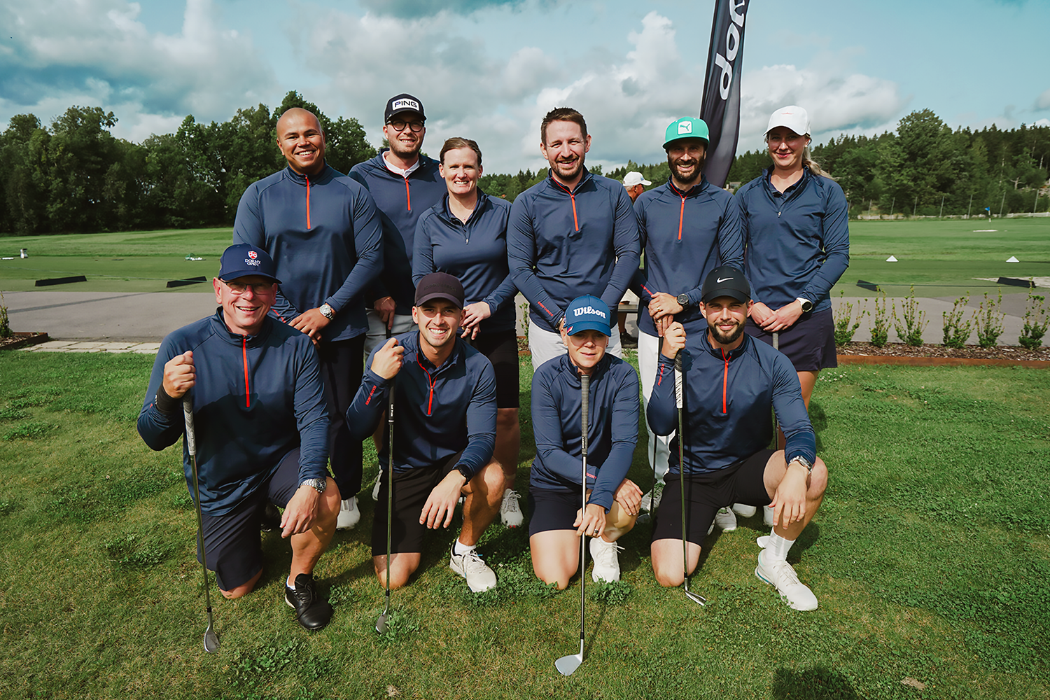 Nio personer i matchande blå golfkläder poserar med golfklubbor på en grön golfbana under molnig himmel.