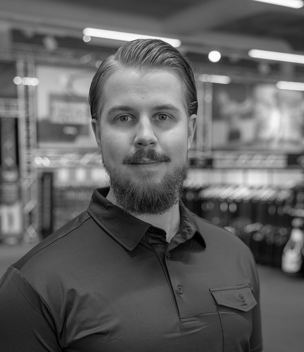 Man med skägg och mustasch står och ler lätt, iförd pikétröja, i en butiksmiljö med suddig bakgrund.