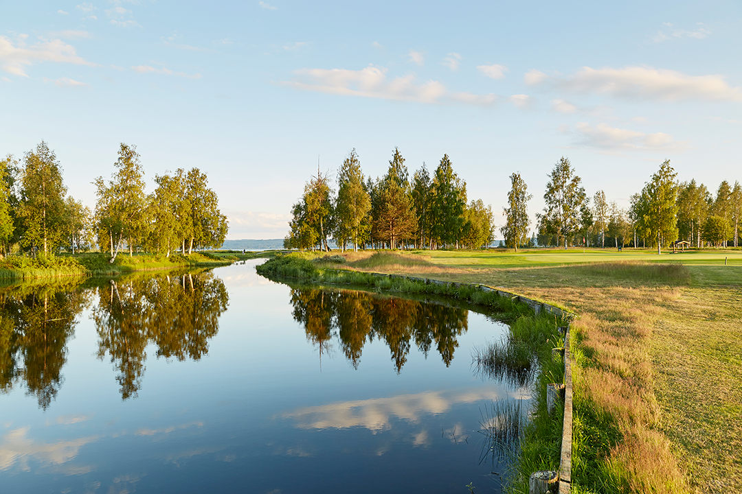En lugn flod speglar träd och himmel, omgiven av gräs och en golfbana i bakgrunden under klar himmel.