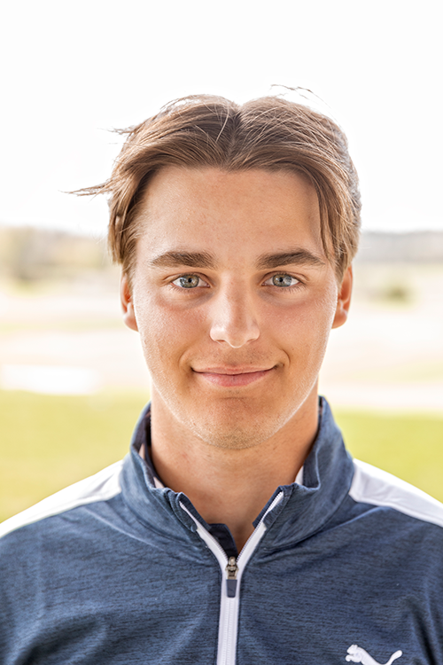 Ung man med brunt hår och blå ögon ler mot kameran, iförd en blå och vit Puma-jacka utomhus i dagsljus.