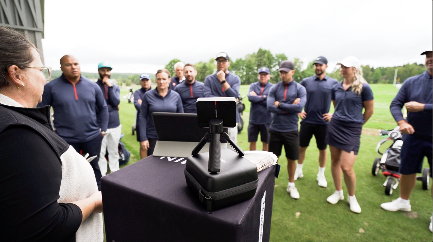 En grupp människor i golfkläder lyssnar på en kvinna som håller en presentation utomhus vid ett bord med teknikprylar.