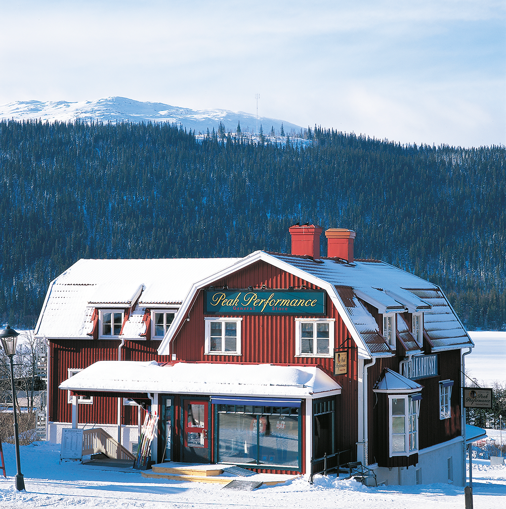 Röd trähus med skylten "Peak Performance General Store", täckt av snö, omgiven av vinterlandskap och skogsklädda berg.
