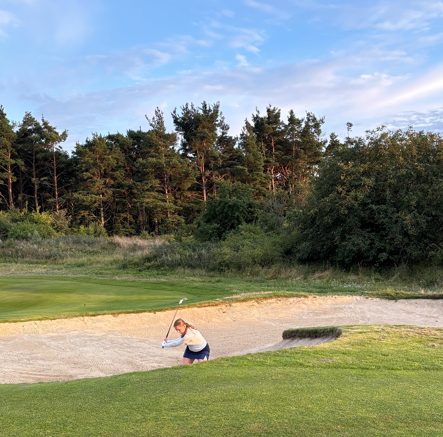 Kvinna slår ett golfslag från en sandbunker på en golfbana med skog i bakgrunden under klar himmel.
