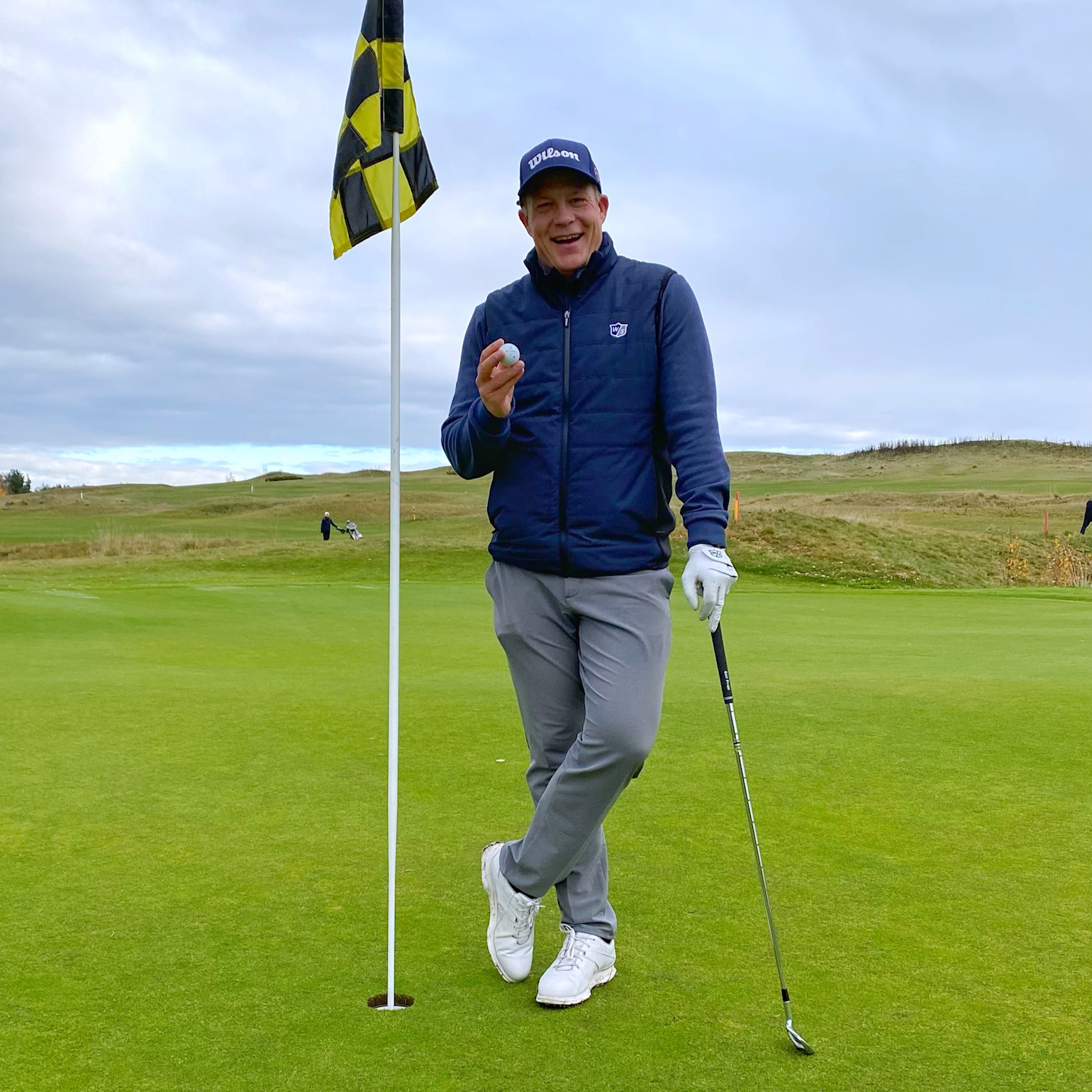 Man smiler og holder en golfbold ved siden af flagstangen på en golfgreen, med en golfkølle i den anden hånd og landskab i baggrunden.