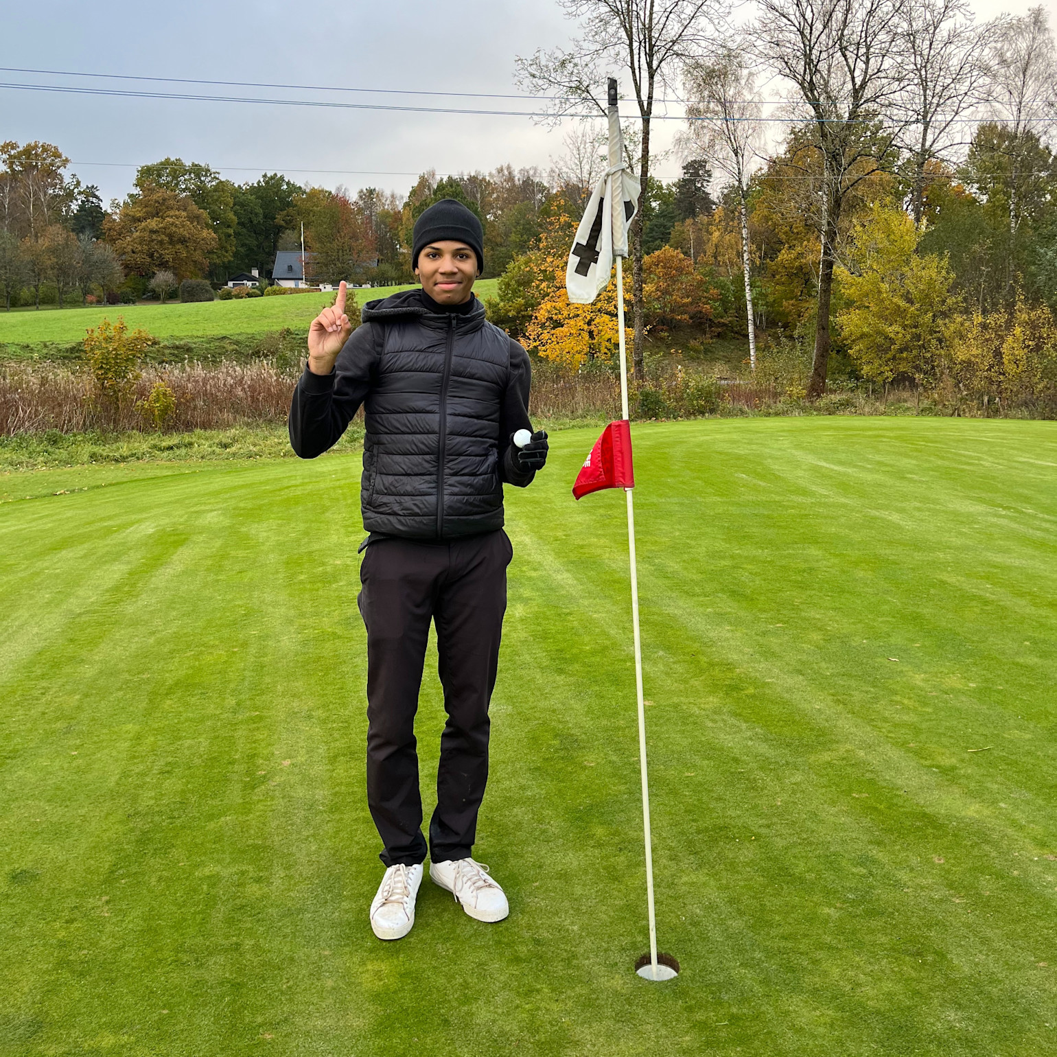 Man i svart jacka och mössa står bredvid en golfhålflagga på en golfbana, håller upp ett finger och en golfboll.