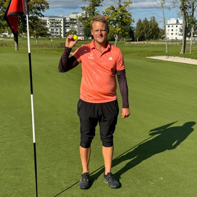 Mies oranssissa pikeepaidassa pitää keltaista golfpalloa kädessään lipputangon vieressä golfkentällä, taustalla rakennuksia.