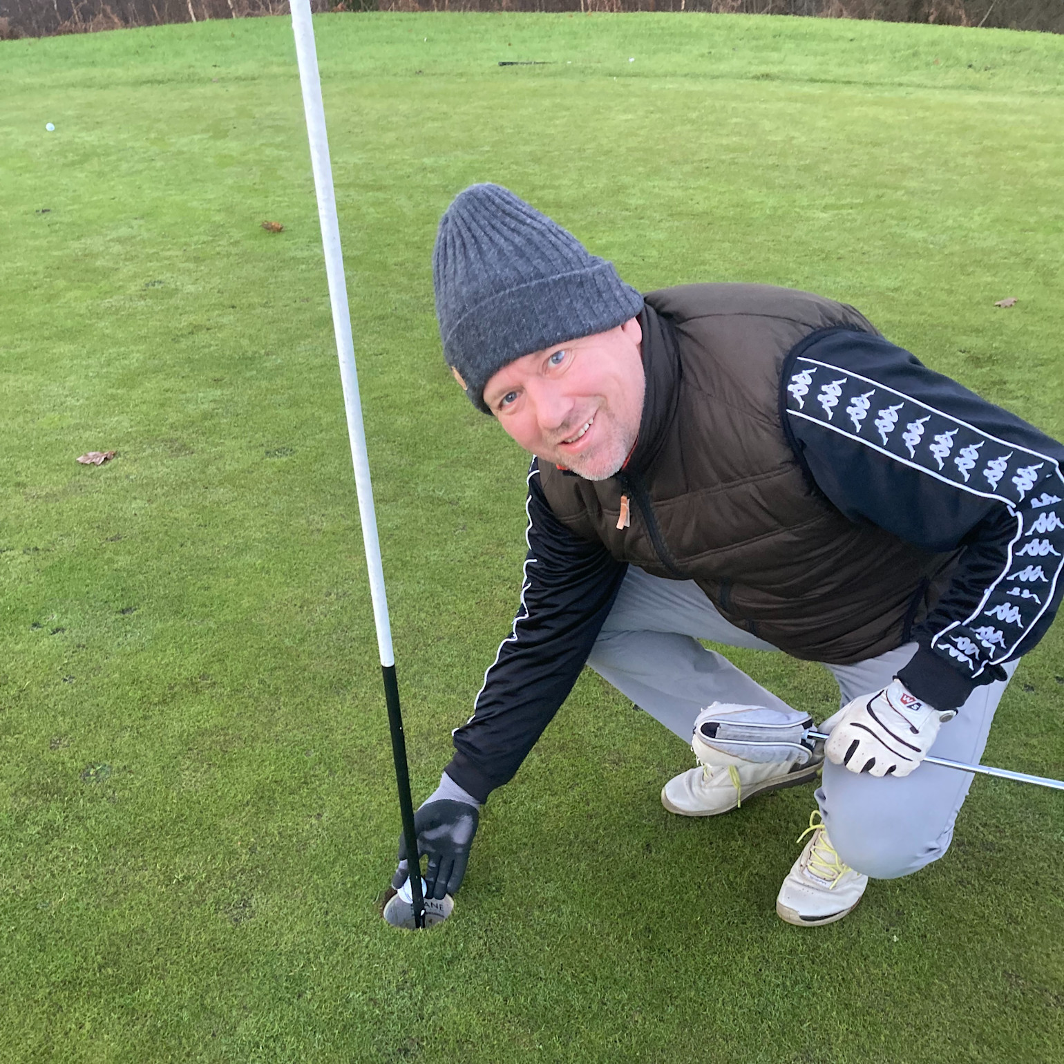 Man i mössa och golfhandske böjer sig ner vid golfhål och plockar upp en golfboll på en green.