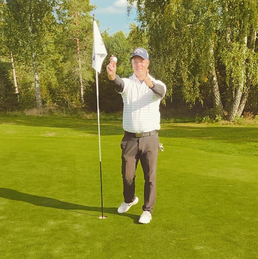 En mand iført golfbeklædning holder en golfbold op og peger mod kameraet ved siden af et hul på en golfbane med grønne omgivelser i baggrunden.
