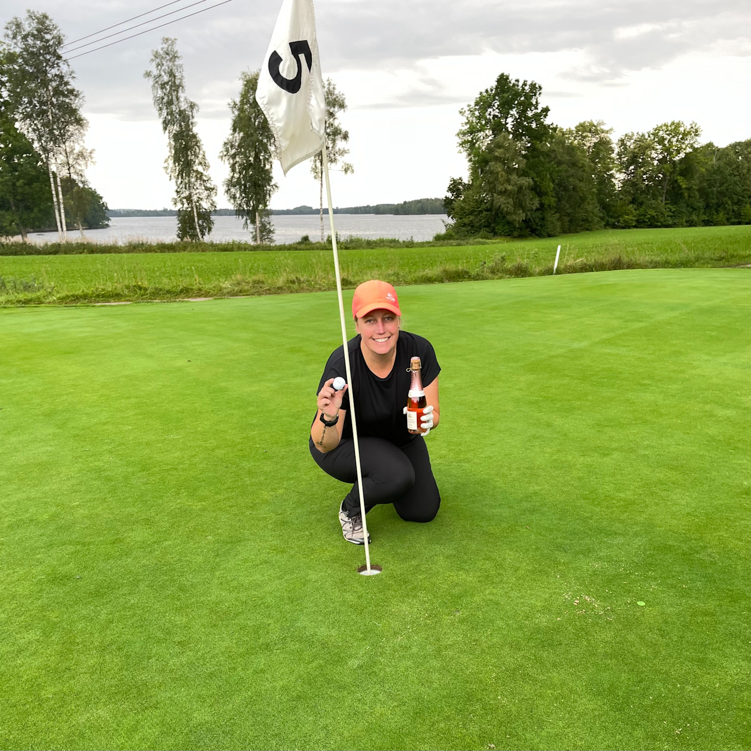 En person knæler ved et golfhul med flag nummer 5, holder en golfbold og en flaske, smilende på en golfbane med en sø og træer i baggrunden.