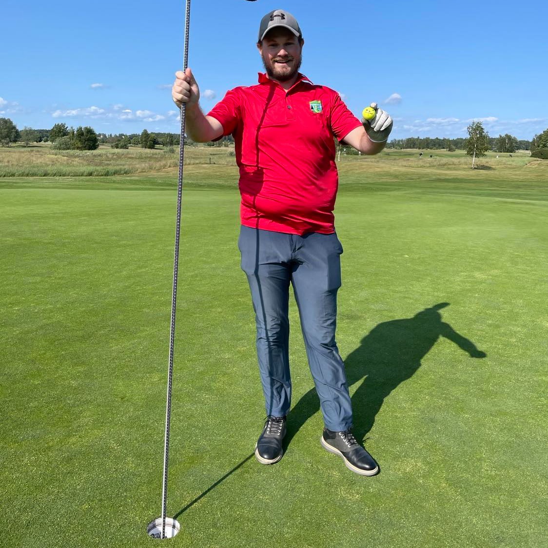 Mand på golfgreen iført rød polo og blå bukser holder en golfkølle og en gul golfbold under en klar blå himmel.