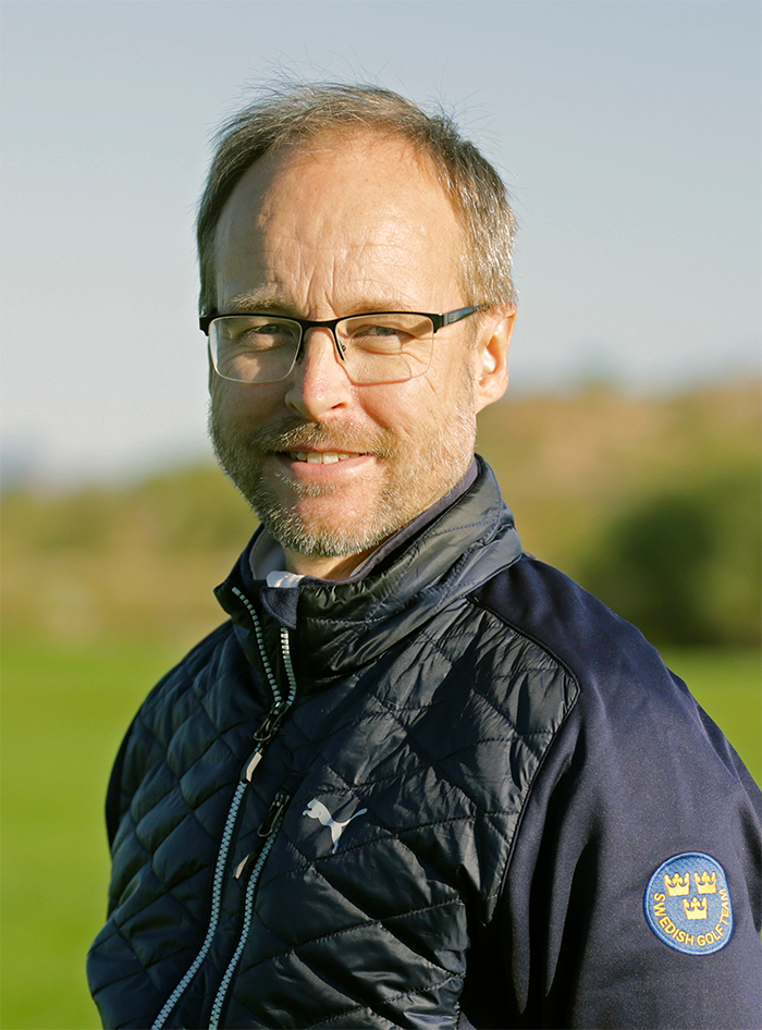 Man med glasögon och skägg bär en mörk jacka med Swedish Golf Team-emblem, står utomhus på en grön gräsmatta.