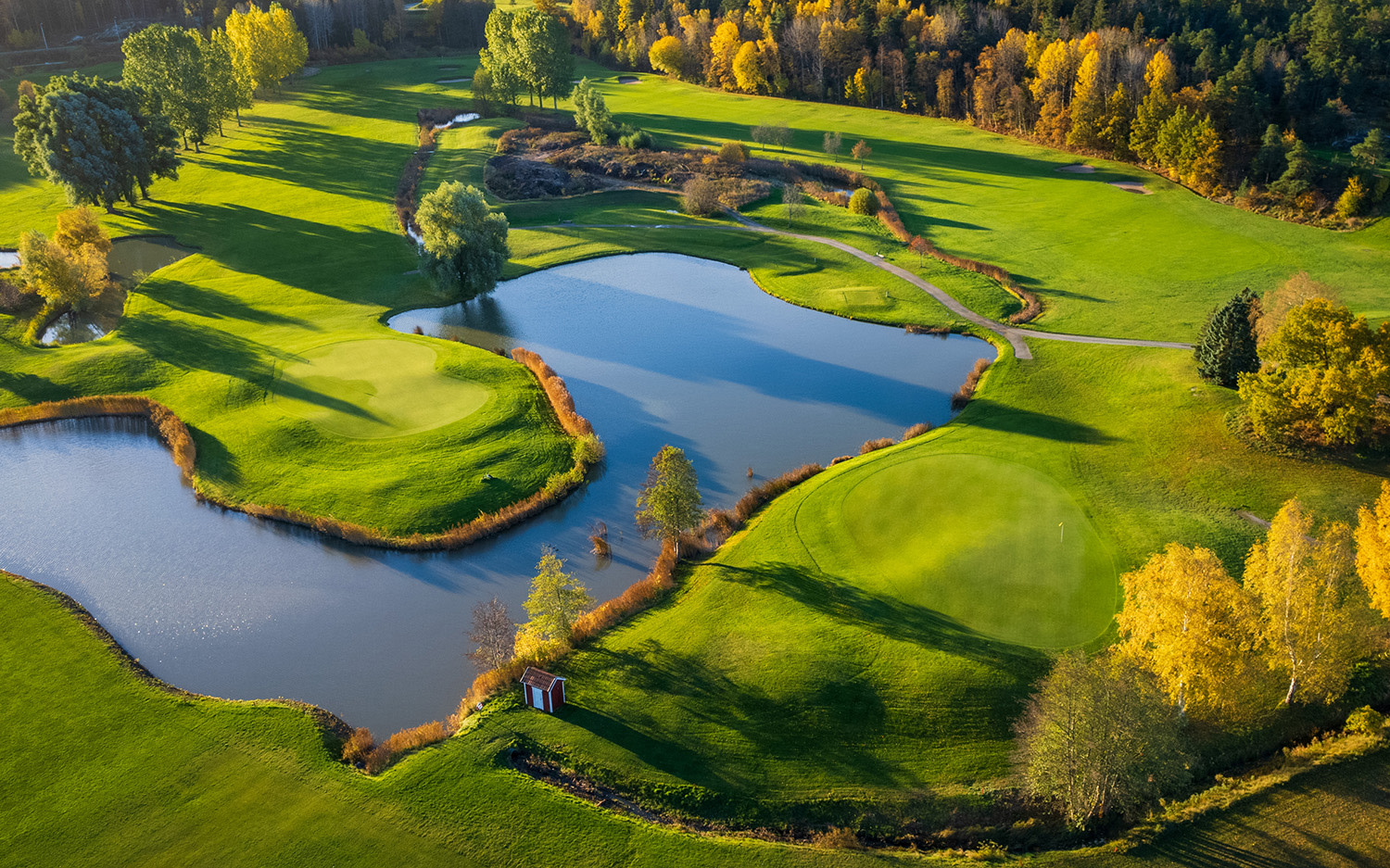 Fågelfotografi av en golfbana med gröna fairways, två dammar, träd i höstfärger och en liten röd stuga vid vattnet.