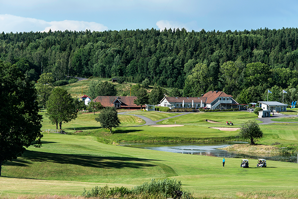 Golfspelare går på en grön golfbana med golfbilar nära en damm, omgivna av träd och byggnader i bakgrunden.