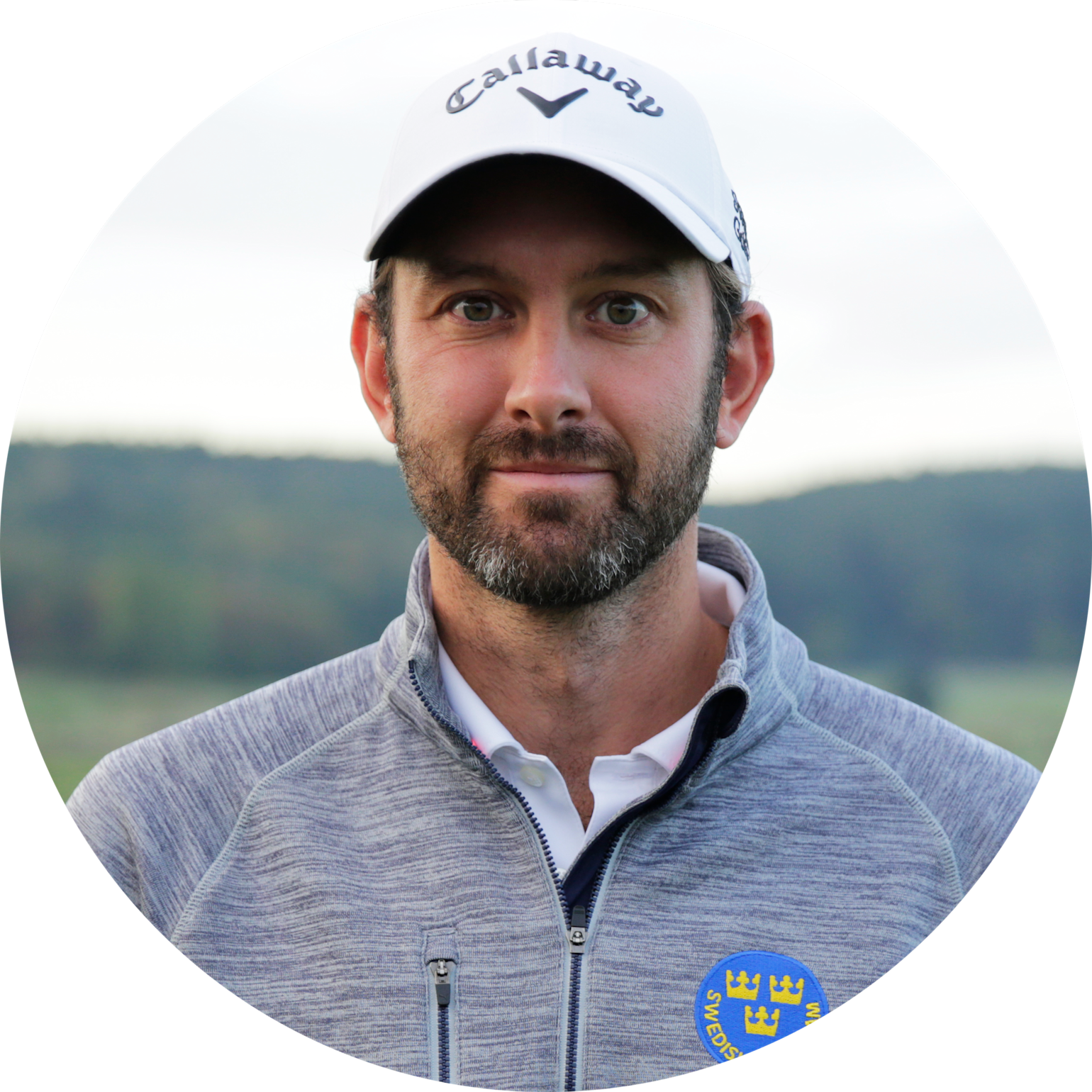 Man med skägg bär vit Callaway-keps och grå jacka med svensk landslagssymbol, står utomhus med suddig natur i bakgrunden.