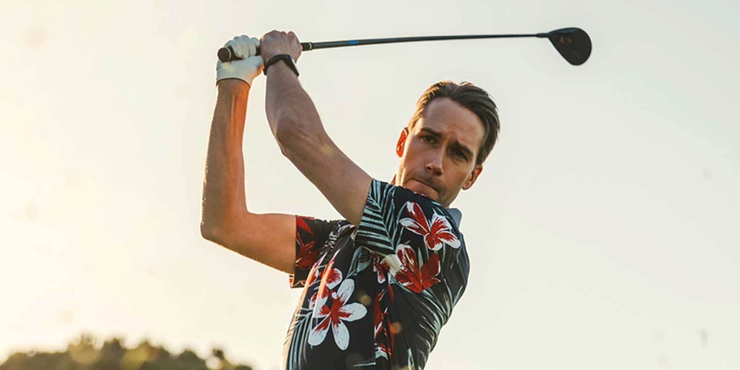 A man is swinging a golf club outdoors, wearing a floral shirt and a golf glove, with a sunset in the background.