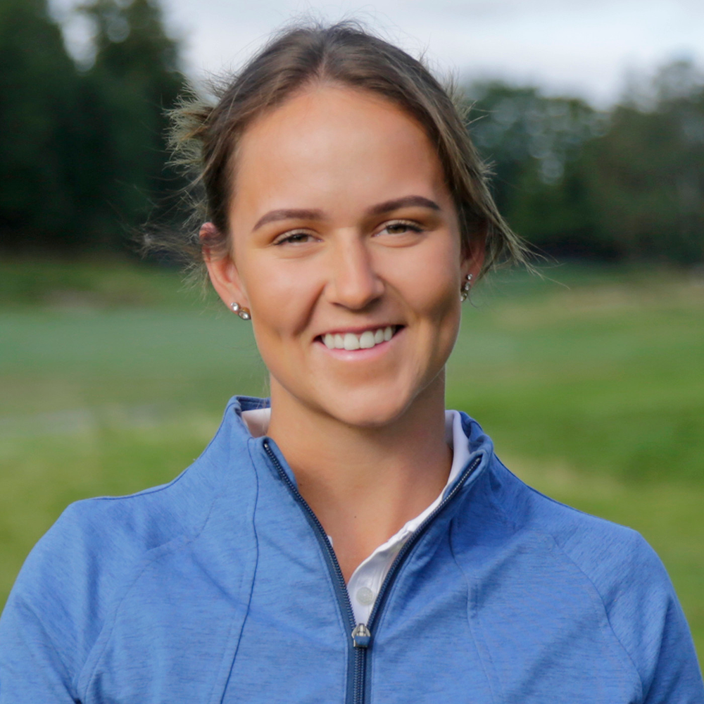 Kvinna med brunt hår och blå jacka ler utomhus på en grön gräsmatta med träd i bakgrunden.