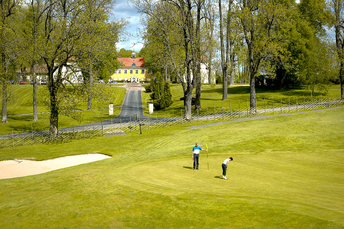 Två personer spelar golf på en grön bana med träd och en gul herrgårdsbyggnad i bakgrunden.