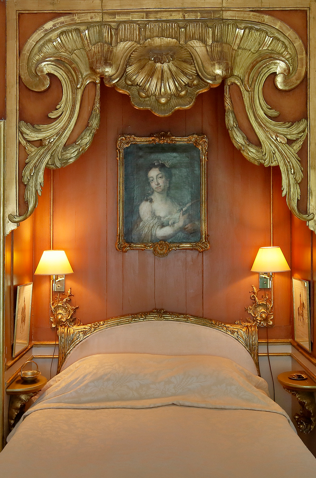 Säng med guldfärgad sänggavel, två vägglampor och ett porträtt ovanför, omgiven av dekorativa gyllene ornament.