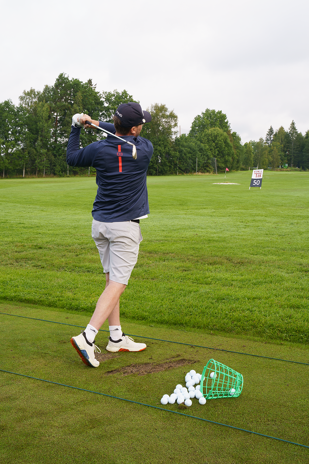 En man svingar en golfklubba på en driving range, med en korg golfbollar på marken och gröna träd i bakgrunden.