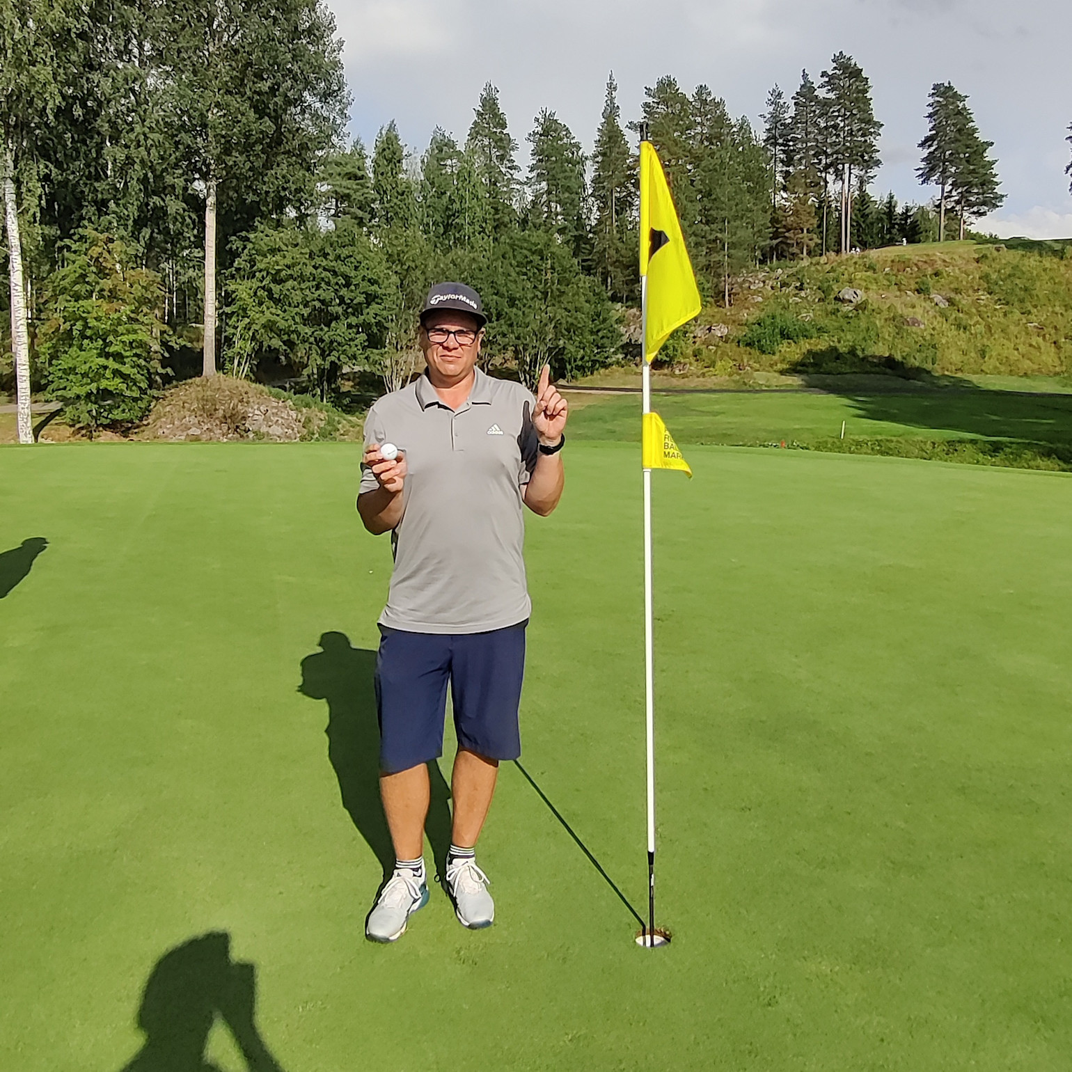 En mand iført golfbeklædning holder en golfbold op og peger opad ved siden af en flagstang på en golfgreen, omgivet af træer.