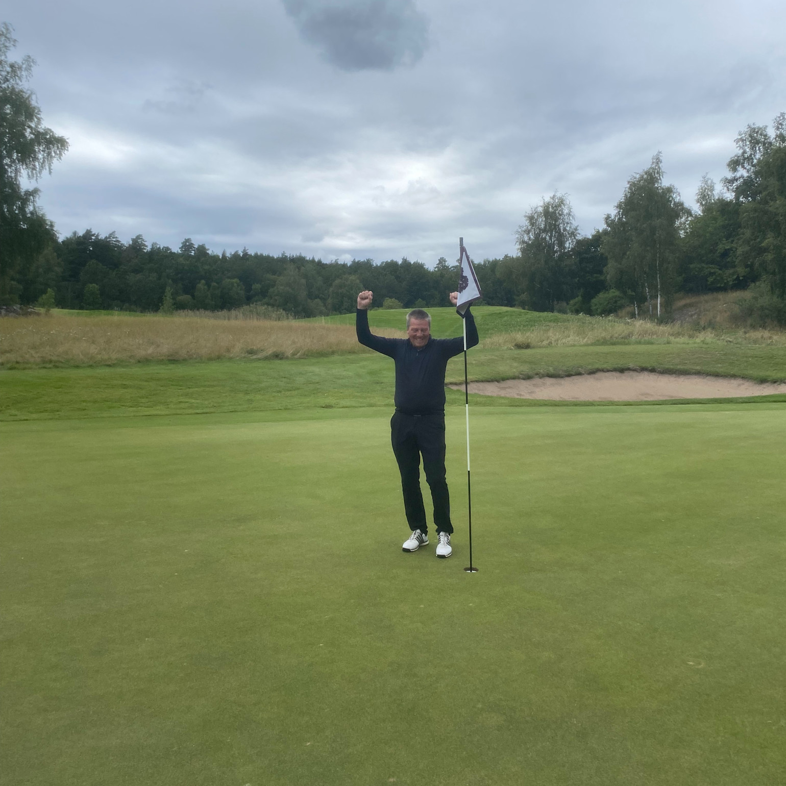 En person fejrer med armene løftet op ved siden af en flagstang på en golfgreen, omgivet af græs og træer under en overskyet himmel.