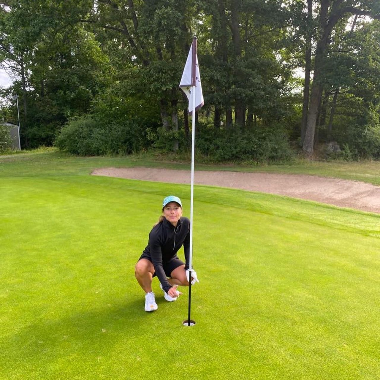 Kvinde i golfbeklædning sidder på hug ved et golfhul og holder i flagstangen på en grøn golfbane med træer i baggrunden.