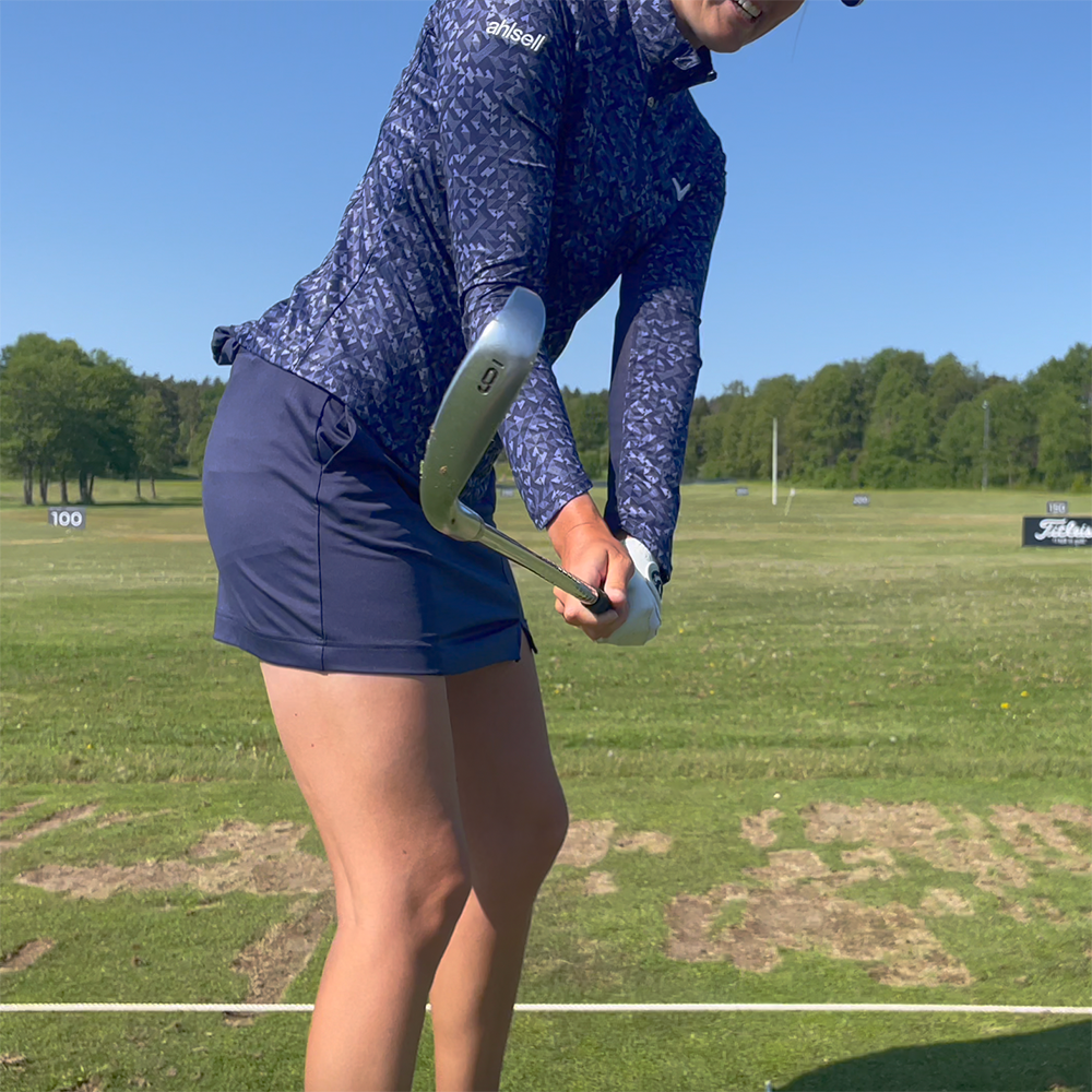 Kvinna i blå golfkläder svingar en järnklubba på en driving range med gräs och träd i bakgrunden.