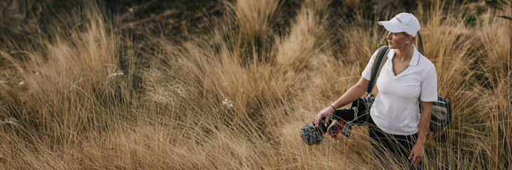 Kvinna bär en golfbag över axeln och går utomhus på en golfbana under en molnig himmel.