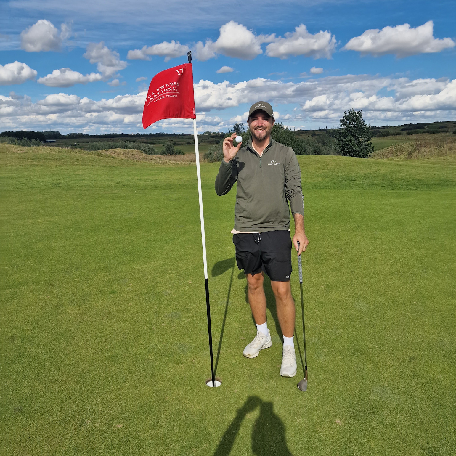 Man holder en golfbold og smiler ved siden af en flagstang på en golfgreen under en blå himmel med skyer.