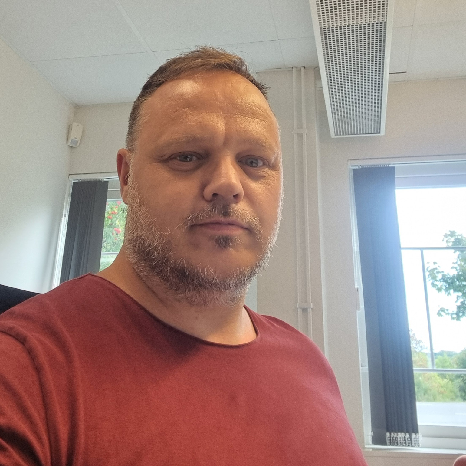 Man med kort hår och skägg bär en vinröd tröja och sitter i ett ljust kontor med stora fönster i bakgrunden.