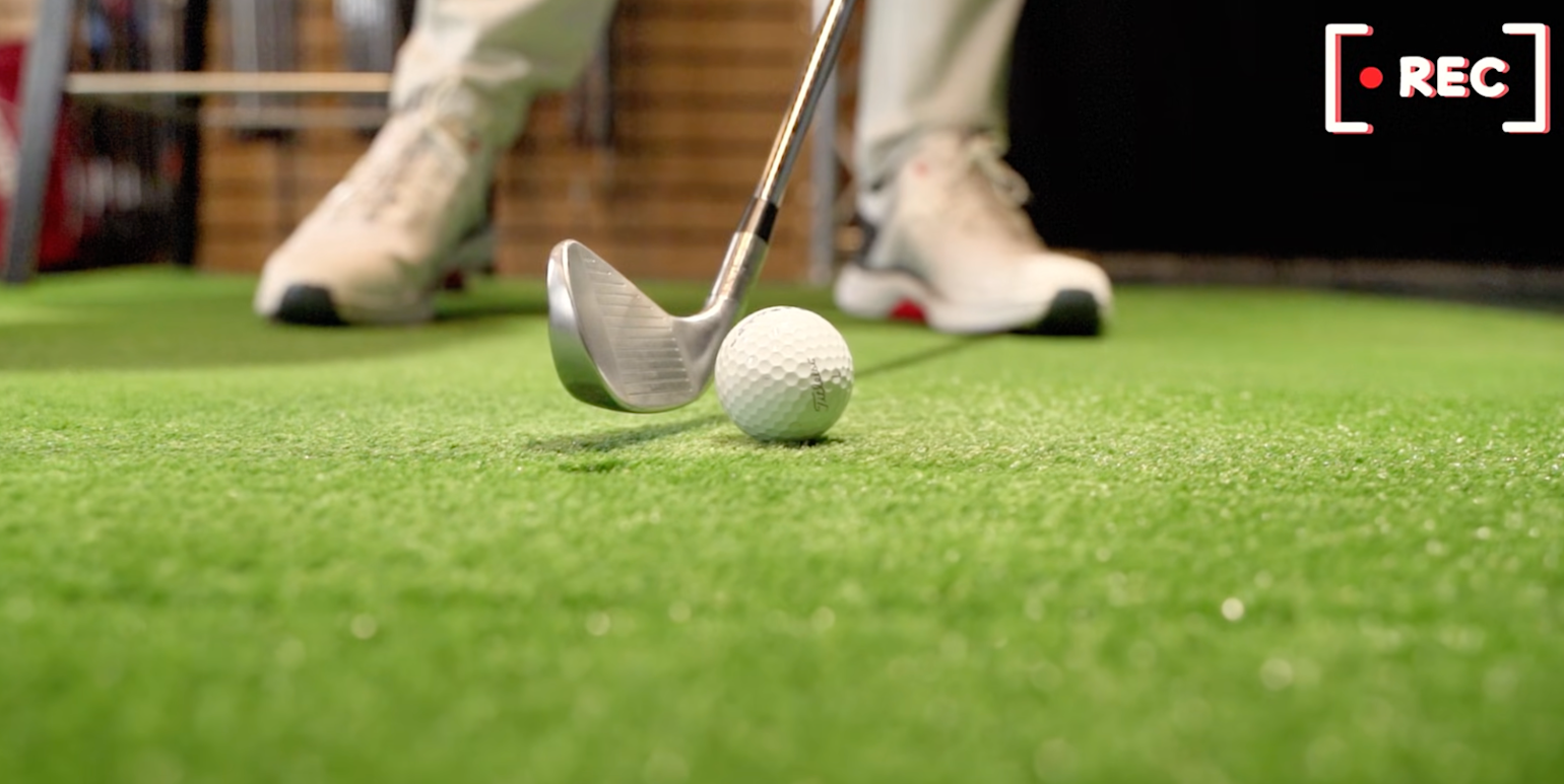 A golf club is aimed at a golf ball on artificial grass, with a person's shoes visible in the background and the text "REC" in the top right corner.