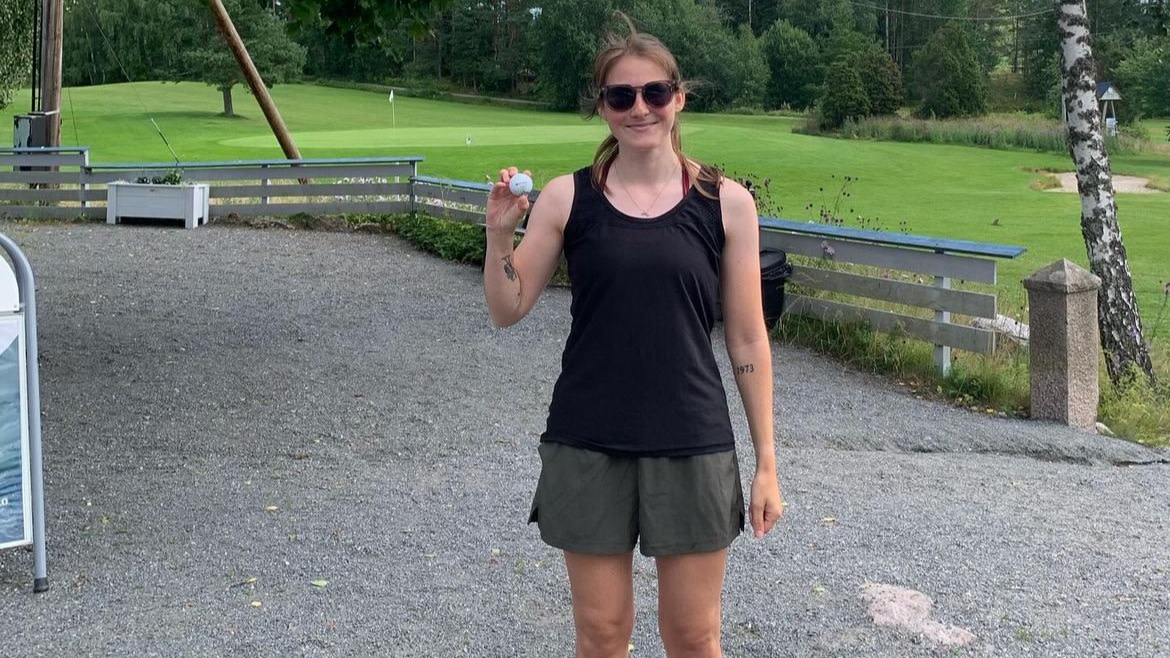 Kvinna i solglasögon håller upp en golfboll och ler, står på grus framför en golfbana med grönska i bakgrunden.