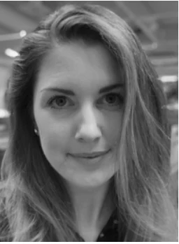 Kvinne med langt hår ser rett mot kameraet i et innemiljø med uskarp bakgrunn.